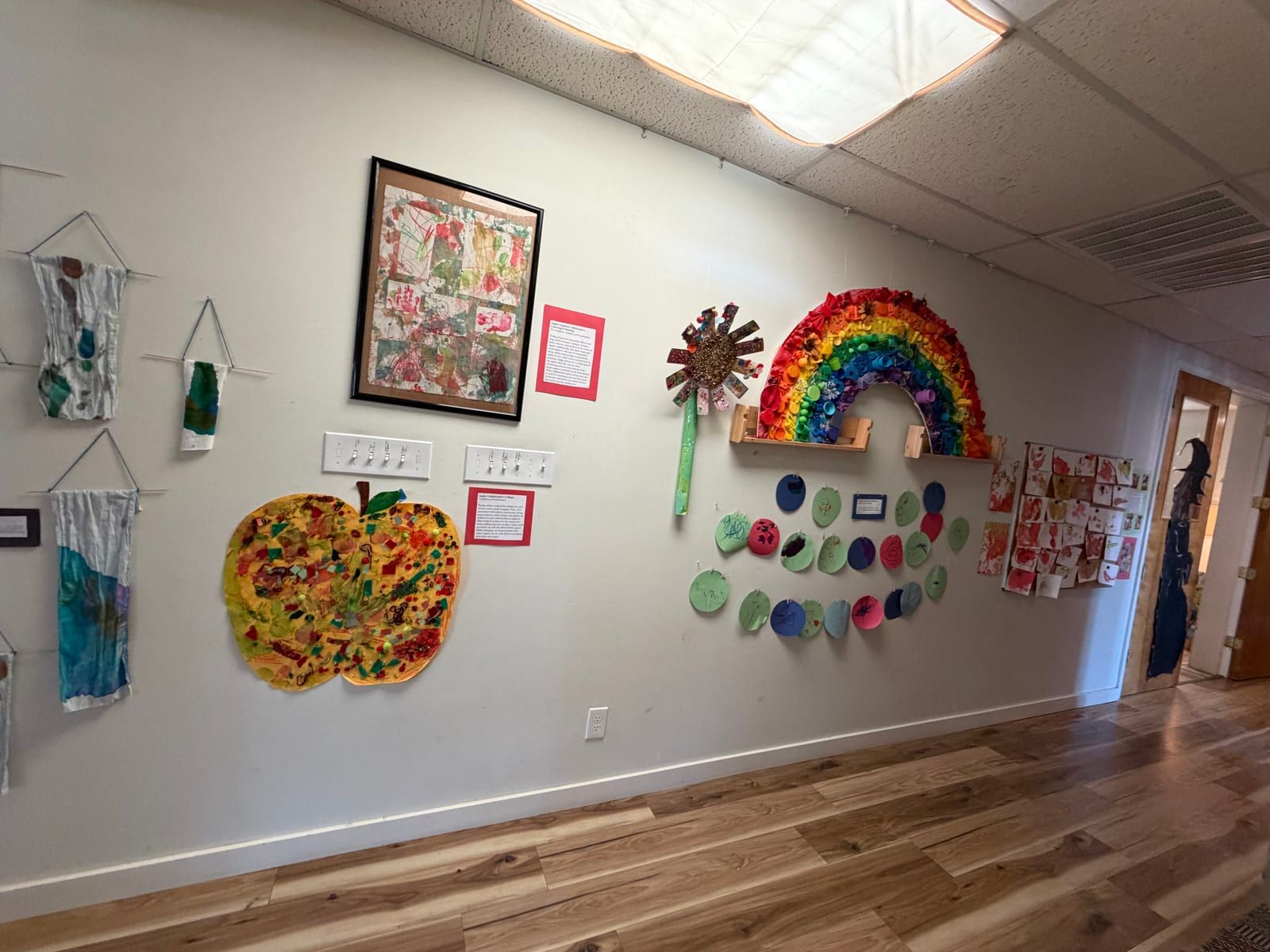A long hallway wall hung with children's art: a rainbow, a big painted apple, fabric pieces and painted plates.