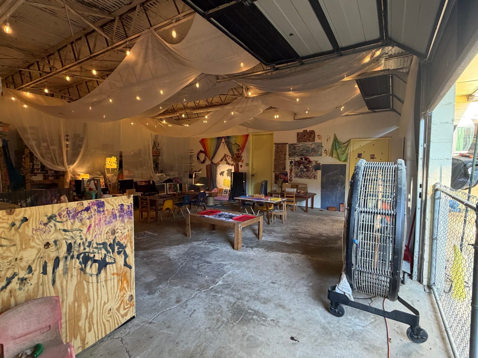 The art studio under draped cloth and string lights, with a painted wooden board in front and tables set with materials.