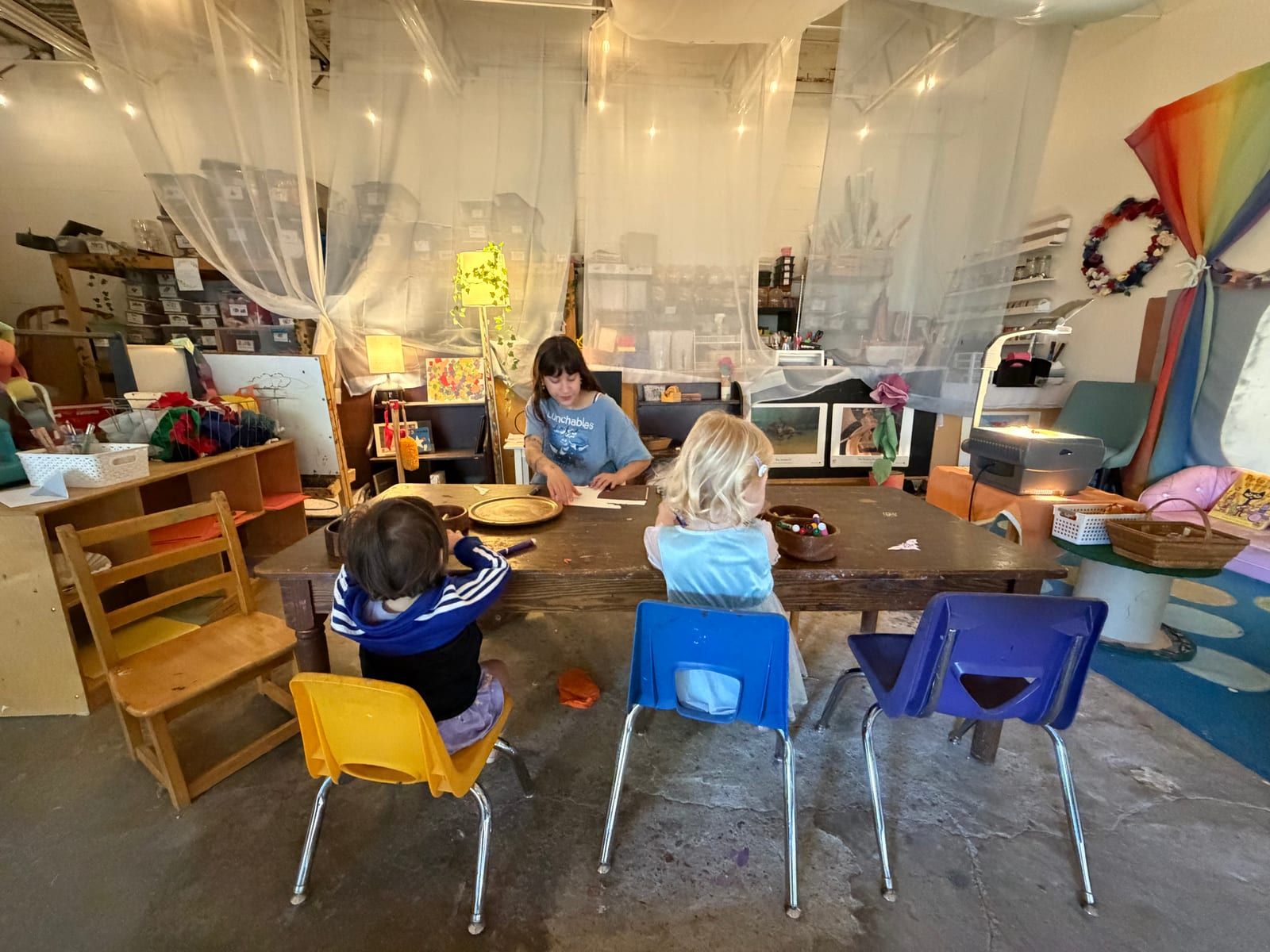 Children on colorful chairs working at a long table in the art studio with a teacher, under sheer draped cloth.