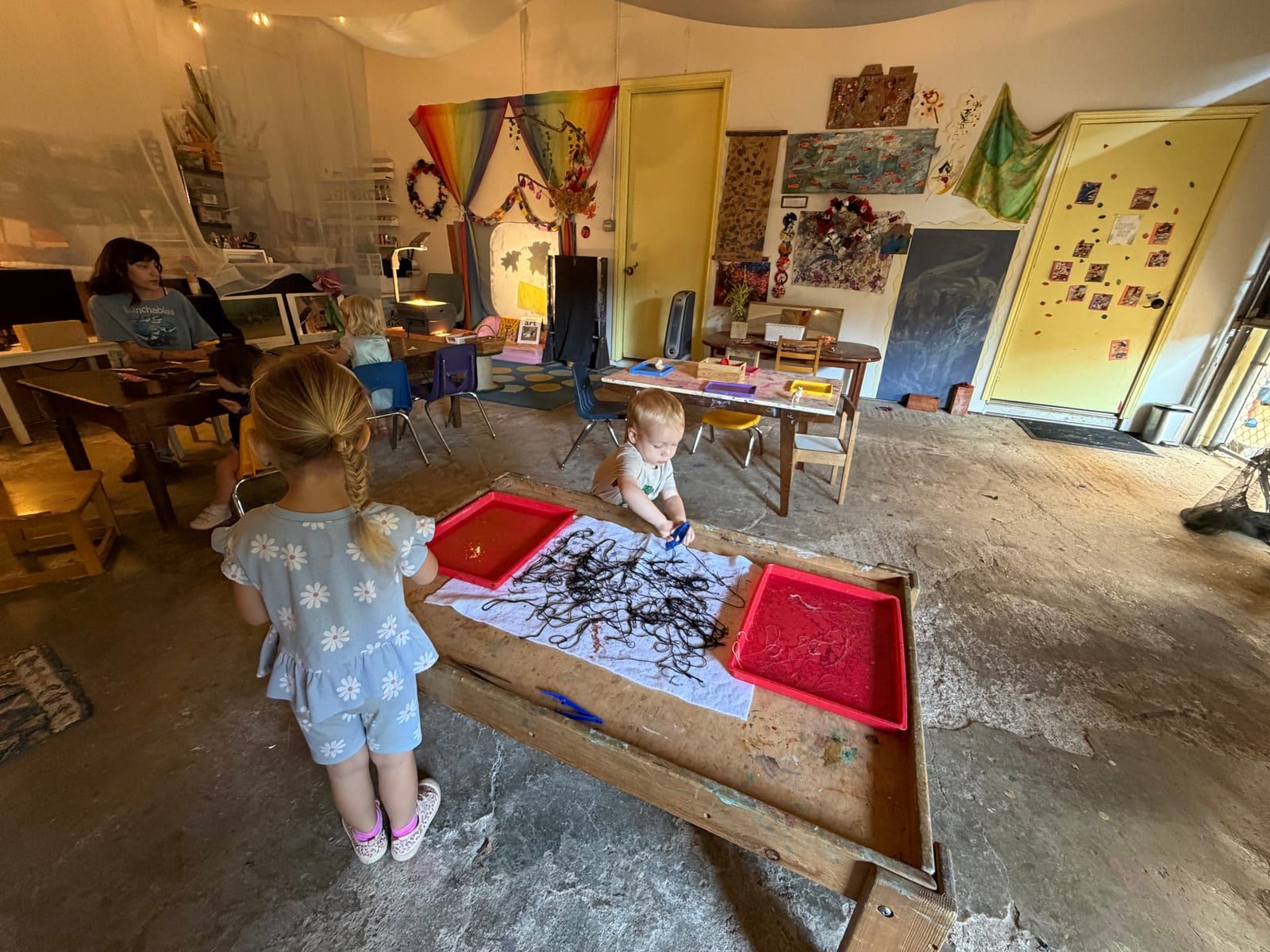 A child painting black lines on paper in a low wooden tray in the art studio, with red paint trays alongside.