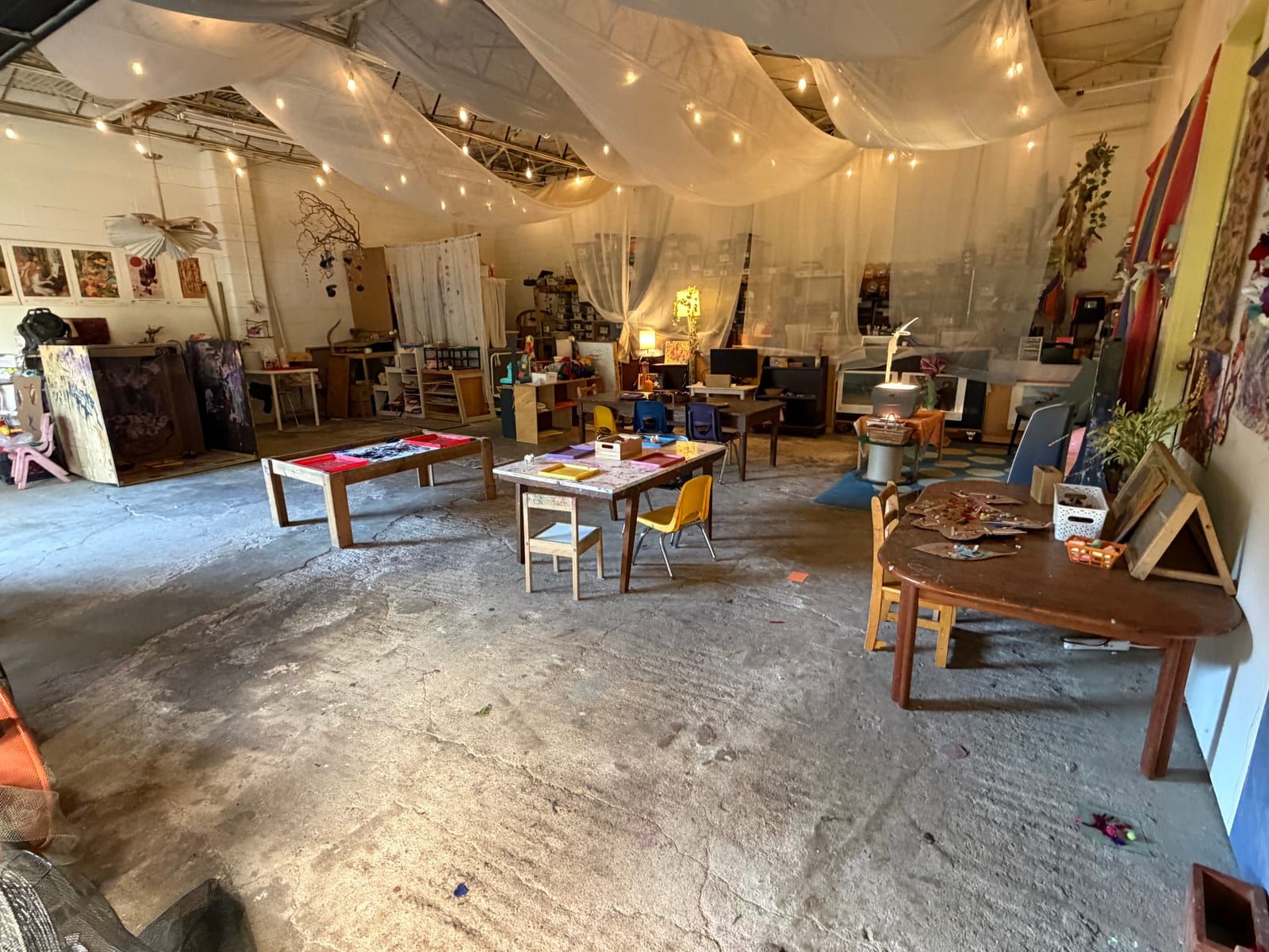 The art studio: white cloth and string lights draped across the ceiling over low tables, small chairs and shelves of materials.