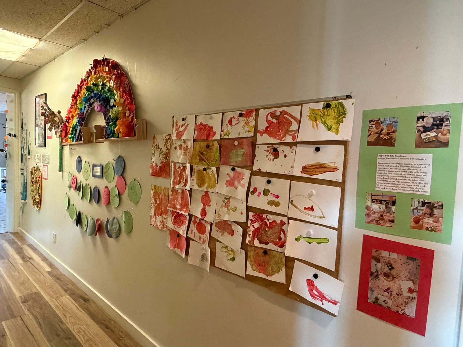 A hallway wall covered in children's work: a rainbow of paper, painted paper plates and rows of red and green paintings.