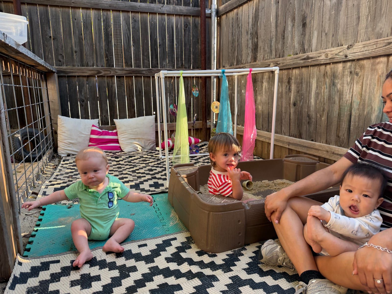 Three babies on an outdoor play mat by a bin of sand and hanging colored fabric, with a teacher holding one of them.