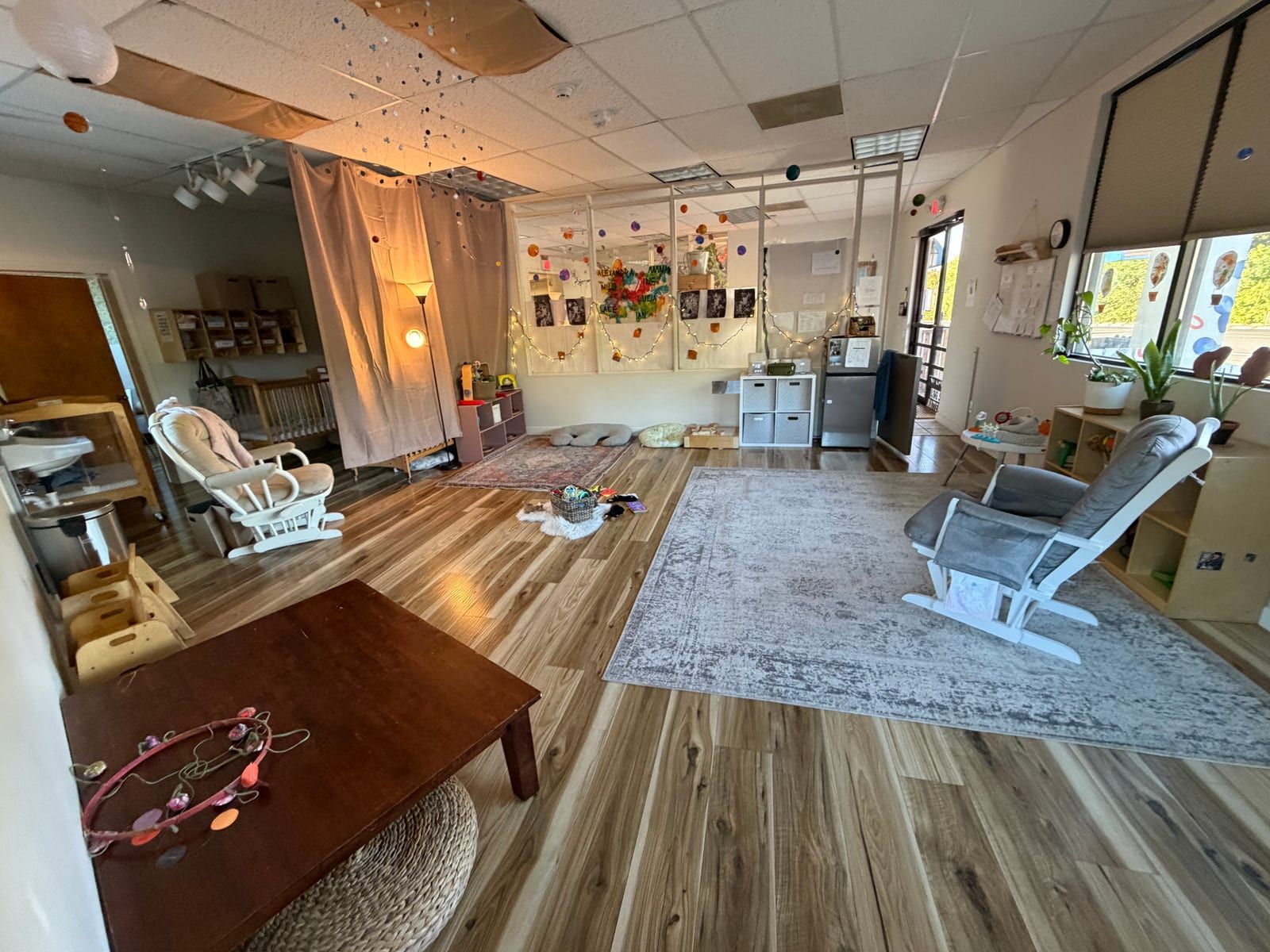 A calm classroom with a wood floor, large rugs, two white rocking chairs, a low wooden table and warm lamps.