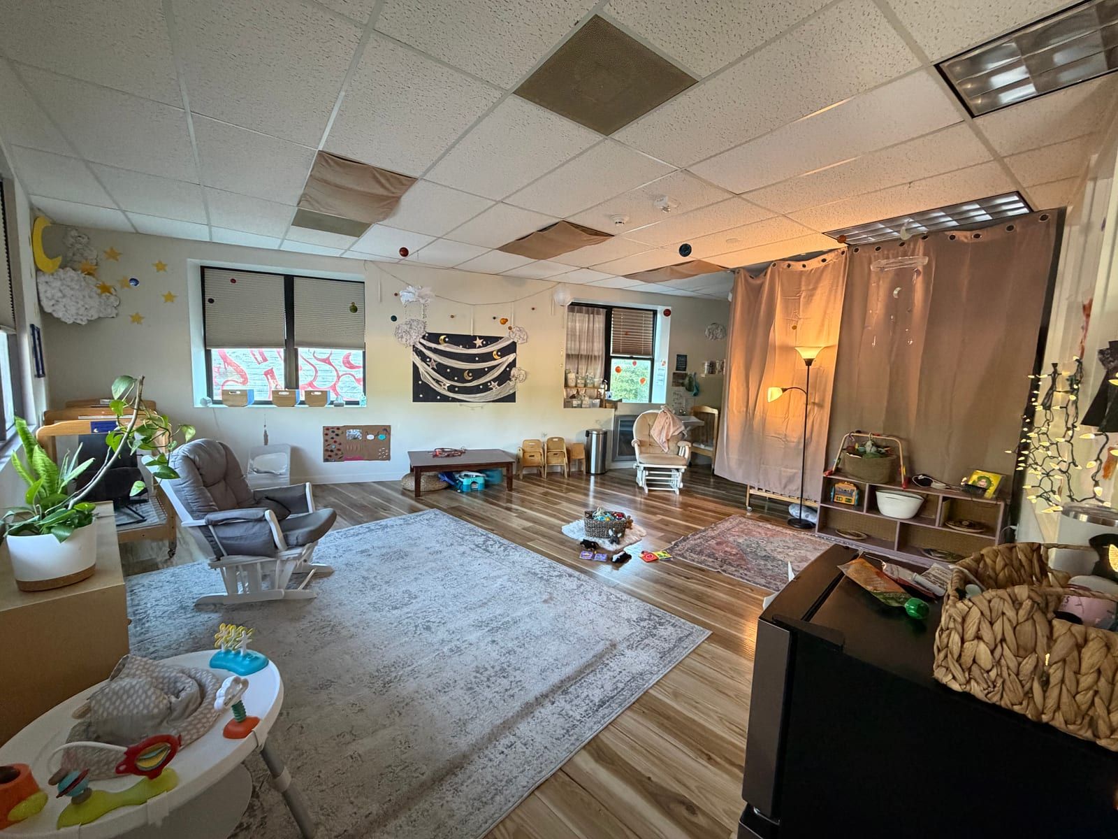A bright classroom with a wood floor, soft rugs, two gliding rocking chairs, plants and low shelves of toys.