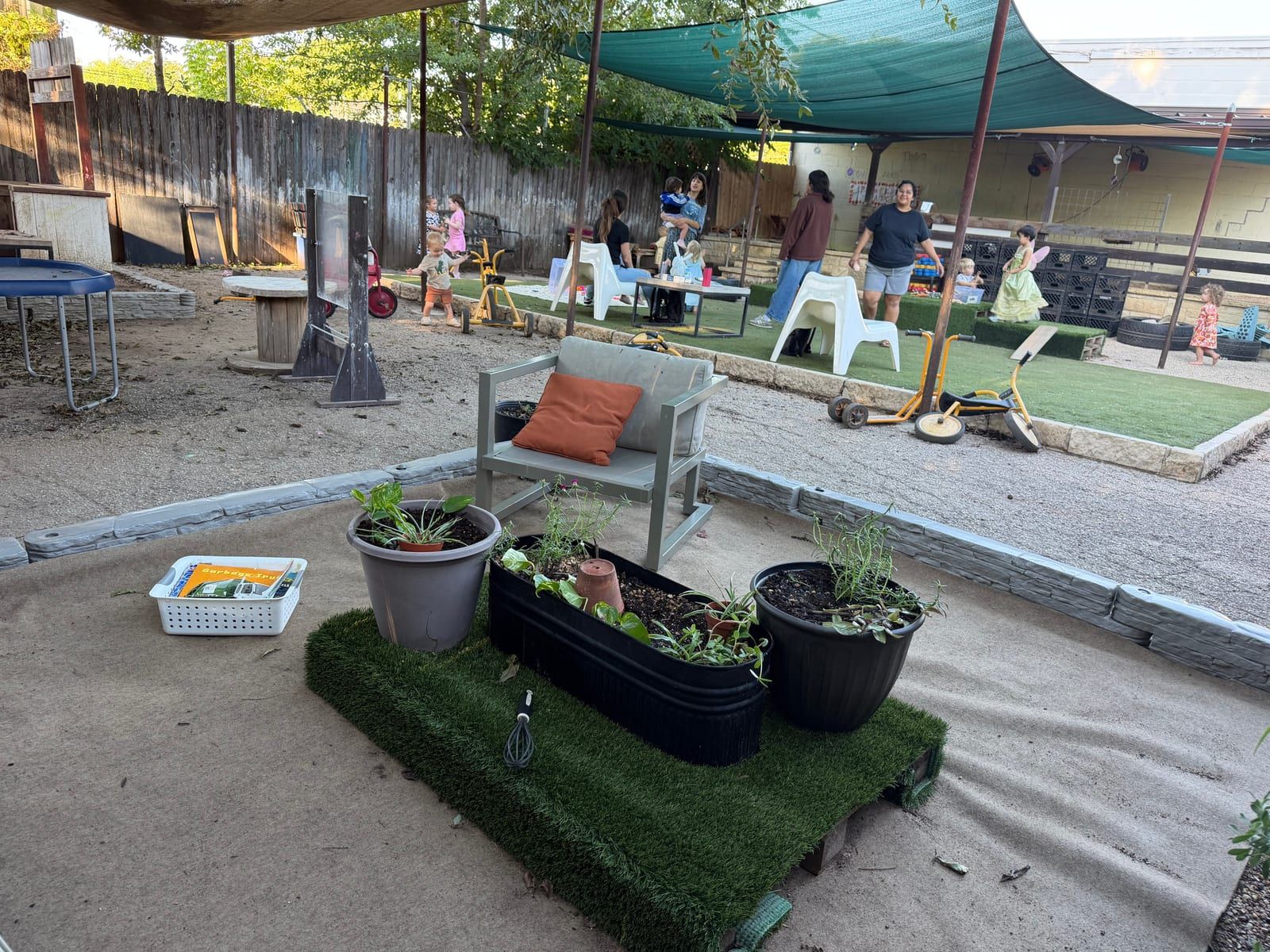 The playground: potted plants on a patch of turf in front, children and parents playing under shade sails beyond.