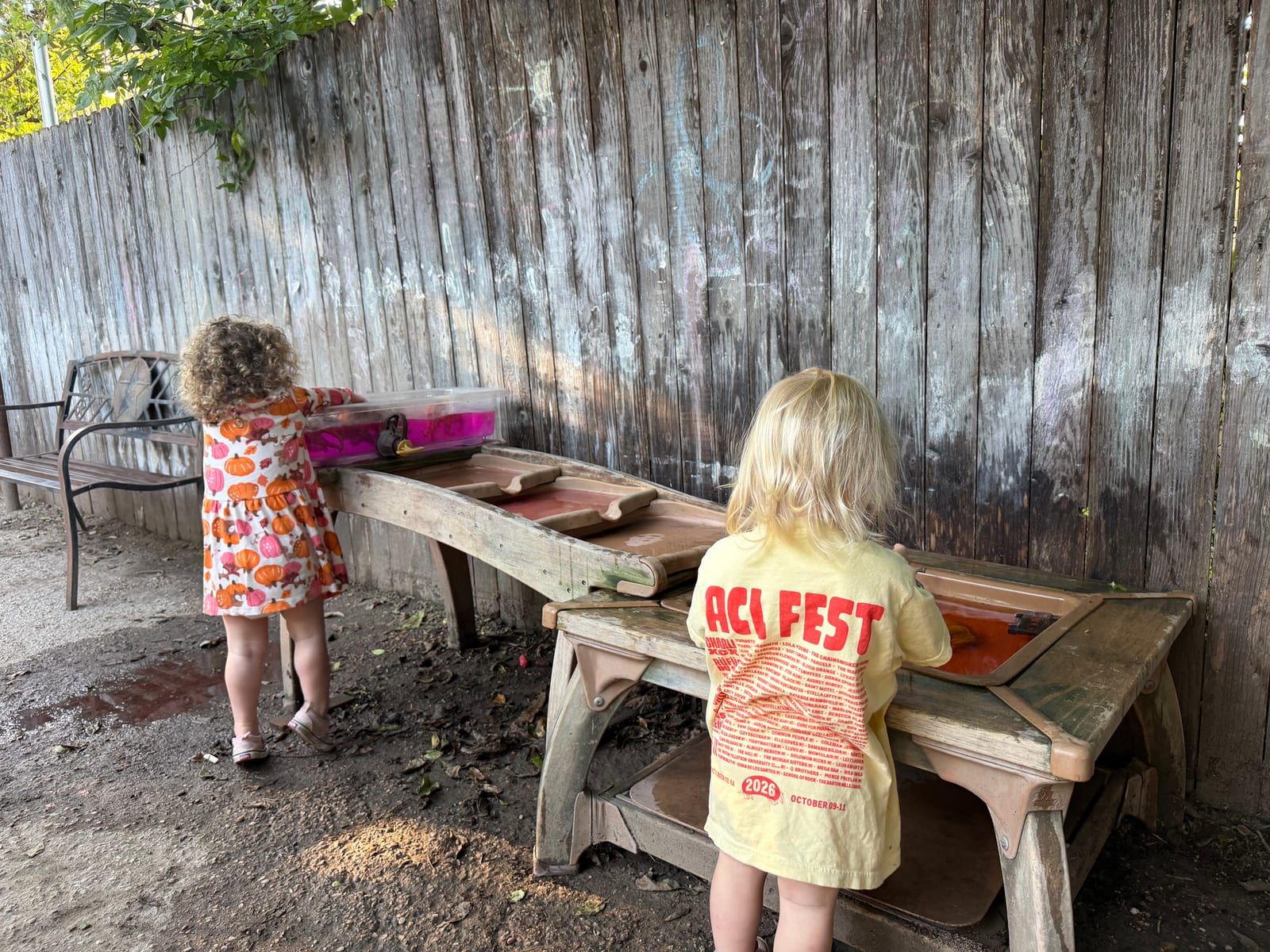 Two young children playing at a wooden outdoor play table, beside a weathered wood fence.