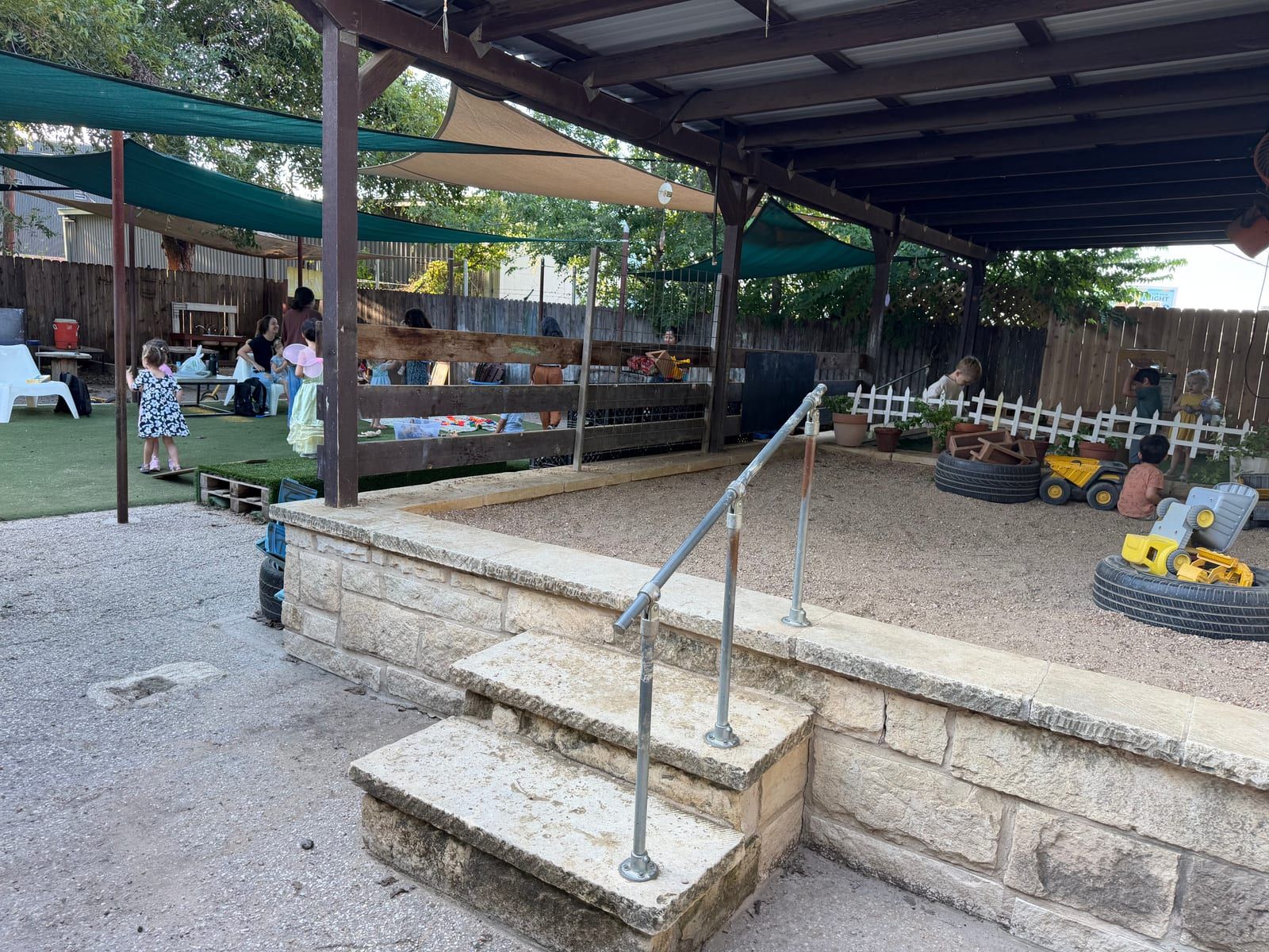 A big sandbox with toy trucks and tires, behind a low stone wall with steps, under a shaded play area.
