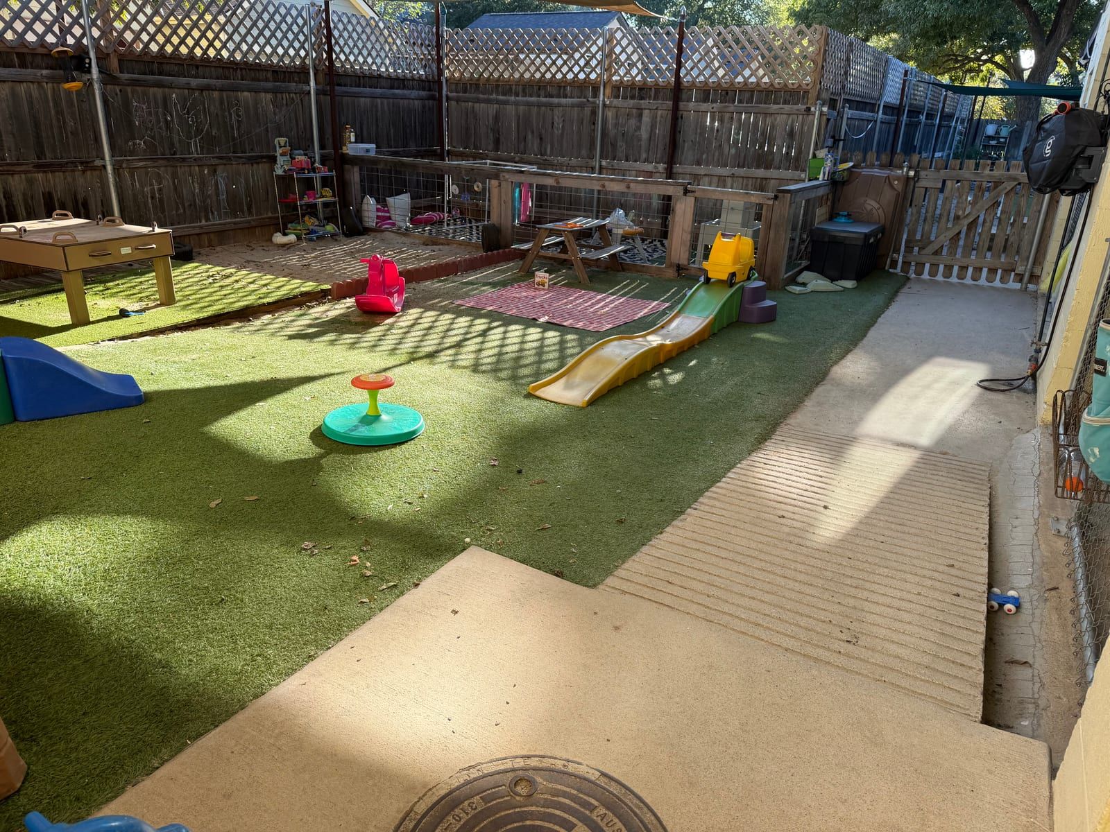 A fenced yard with green turf, a small slide, a spinning seat and a wooden sand table.