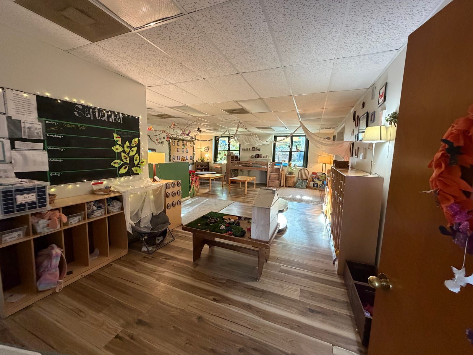 A classroom with a chalkboard, cubbies along the wall, string lights and low wooden tables on a wood floor.
