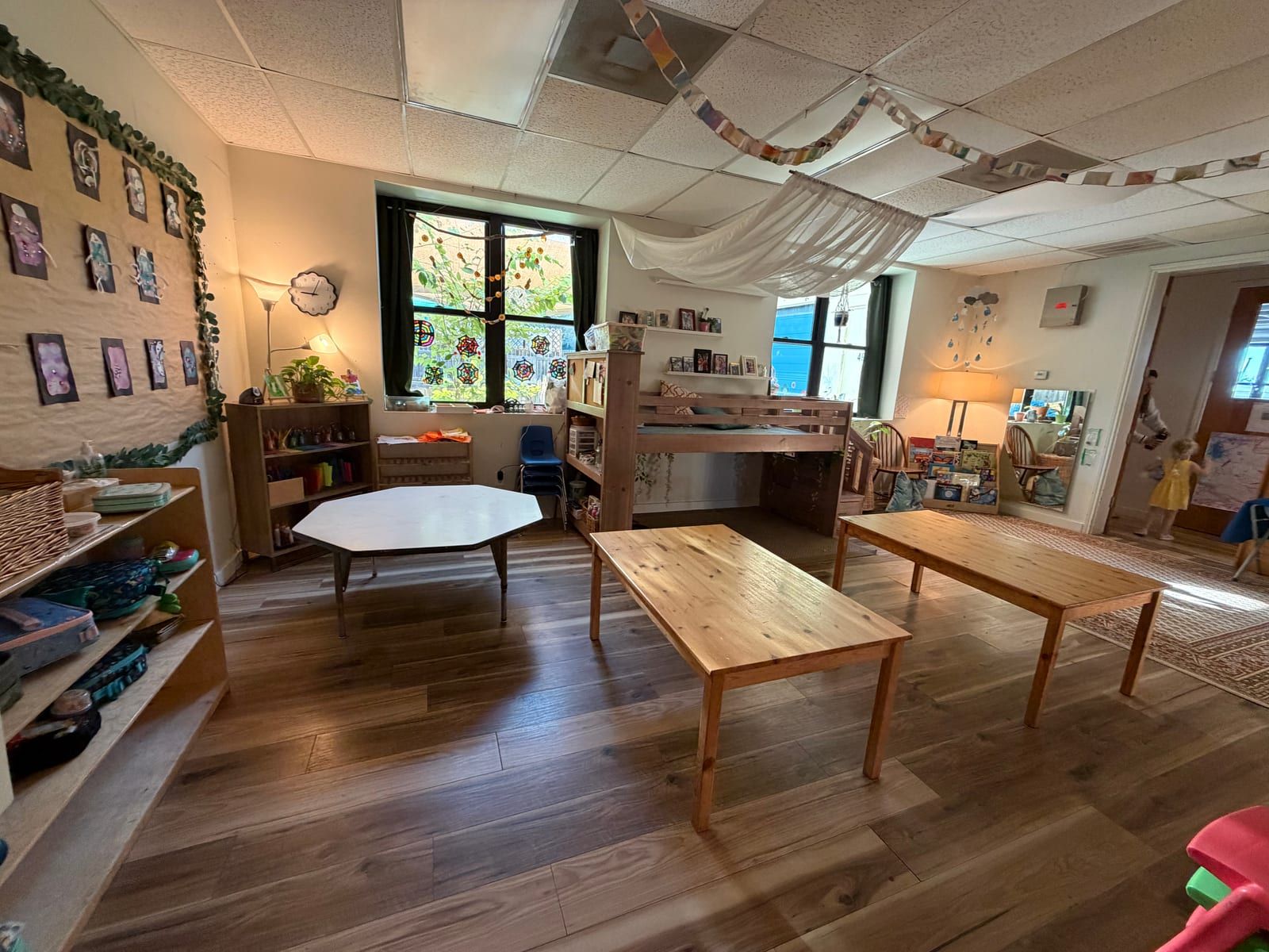A classroom with low wooden tables, shelves of materials, a wooden loft by the windows and framed photos on the wall.