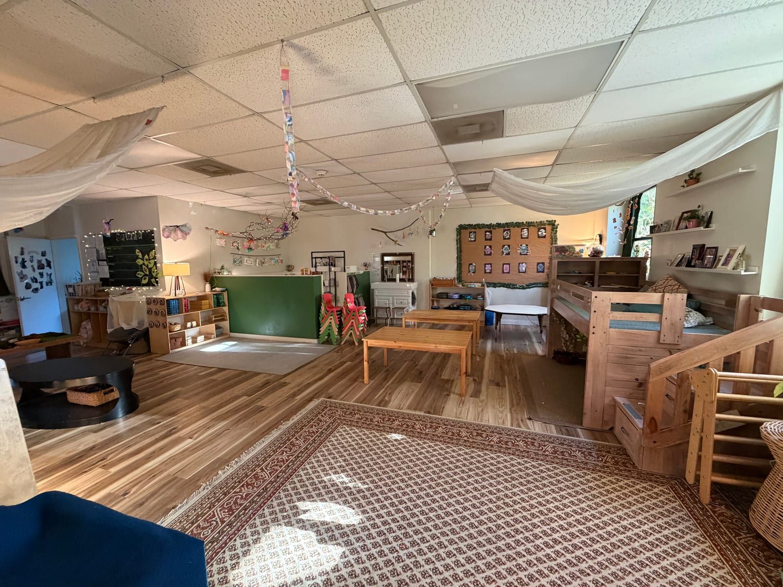 A large classroom with a patterned rug, a wooden loft with stairs, cloth draped from the ceiling and low shelves.