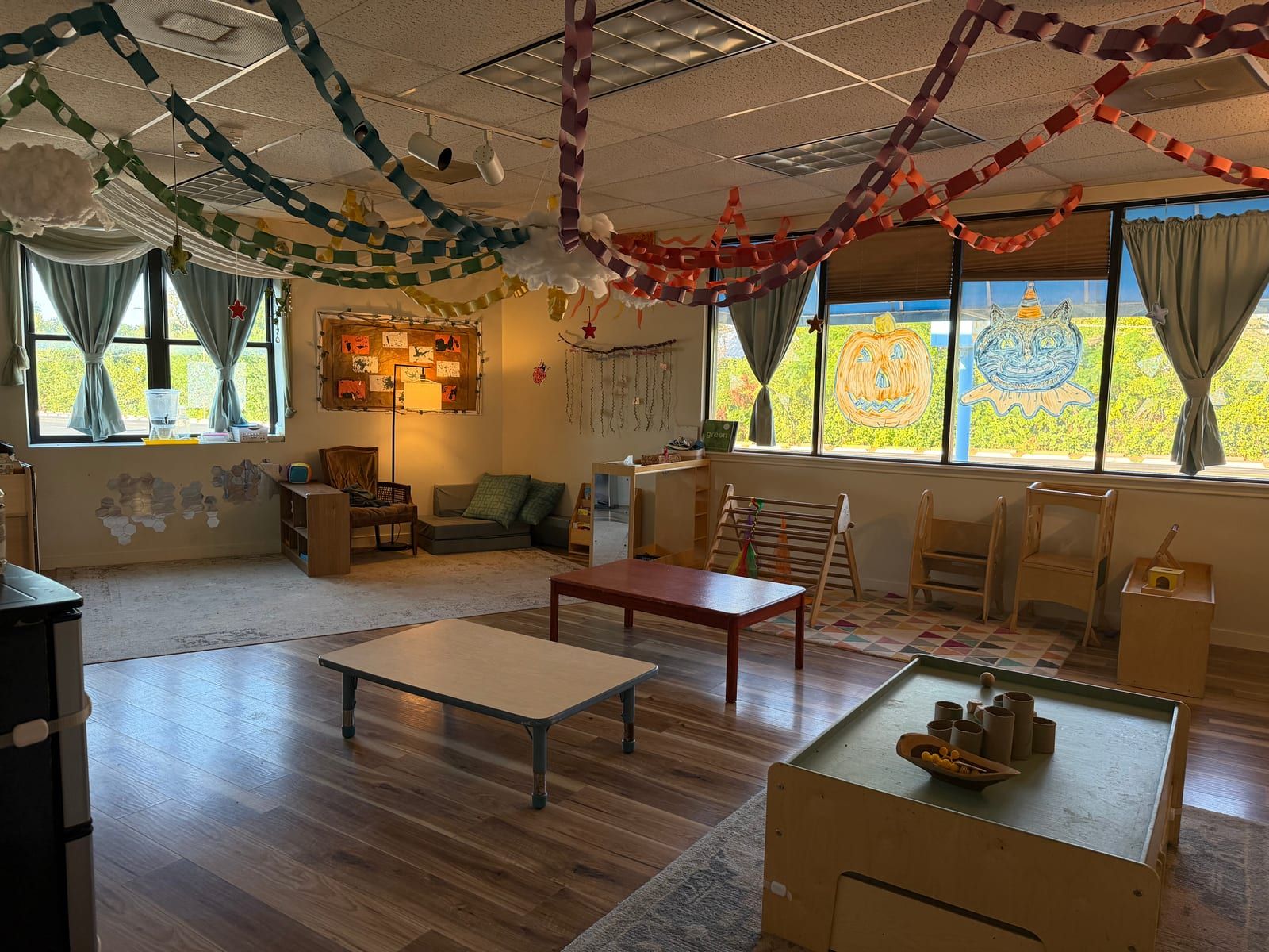 Paper chains strung across a classroom ceiling over low wooden tables, child-sized chairs and windows decorated with art.