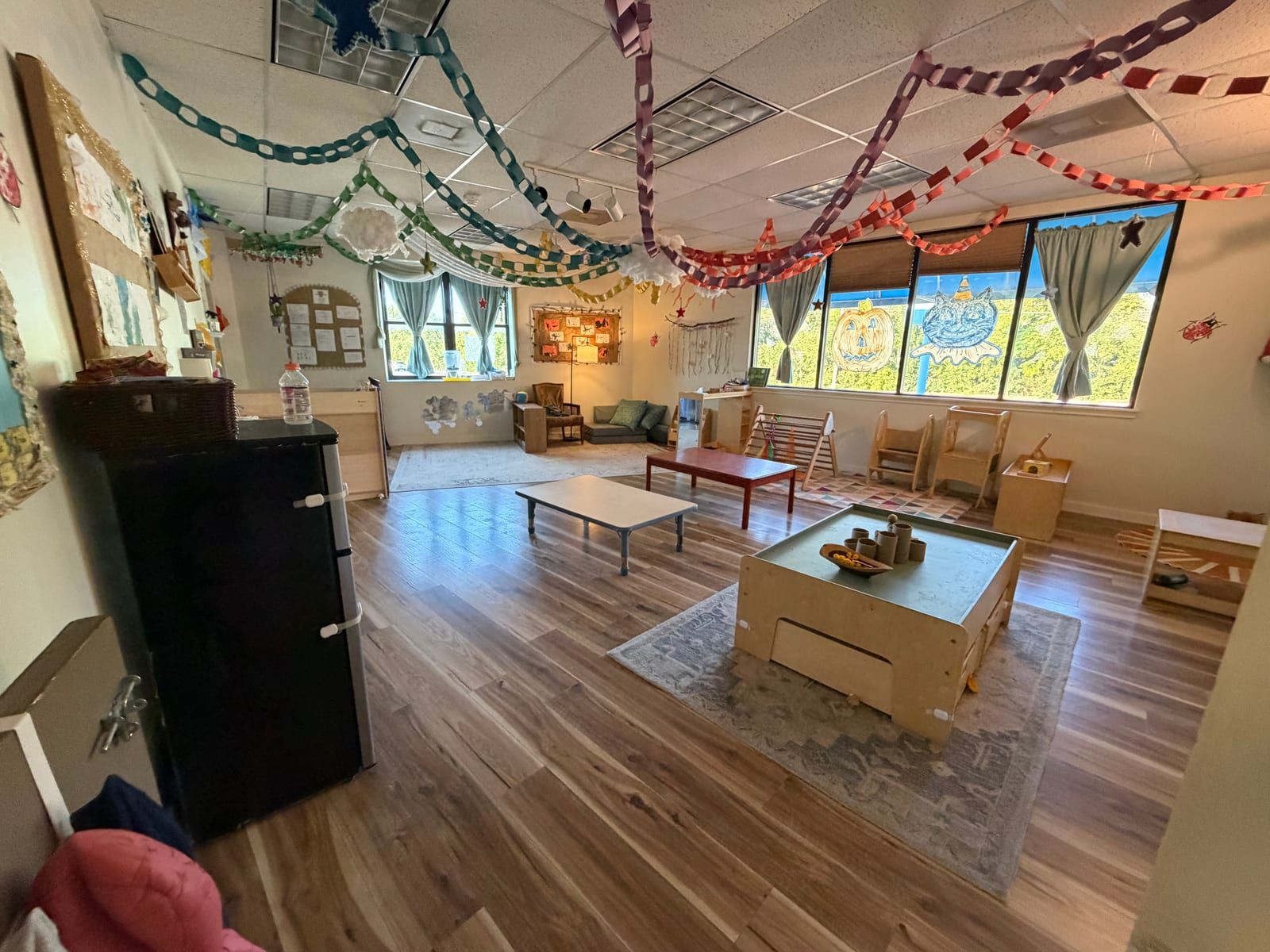 A sunny classroom with paper chains across the ceiling, a wall of windows, low tables and a play table on a rug.