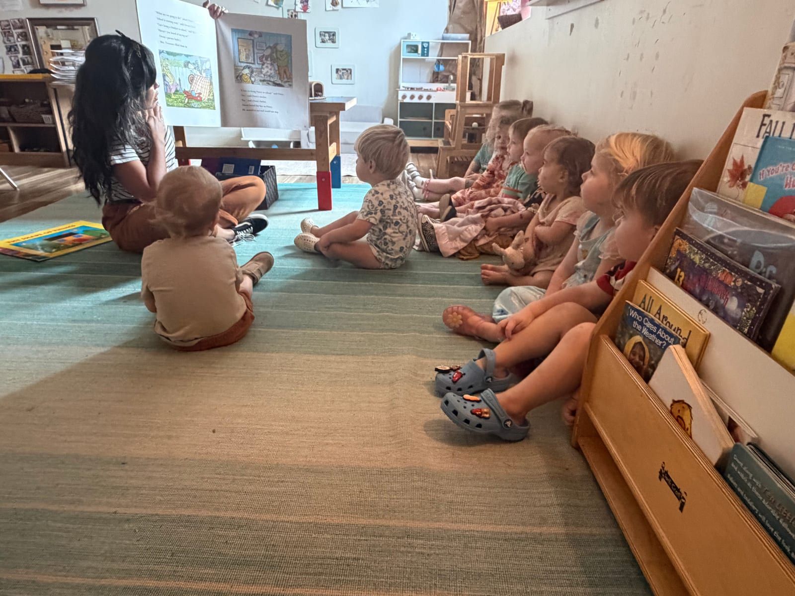 A row of young children sitting on a striped rug beside a bookshelf, listening to a teacher.