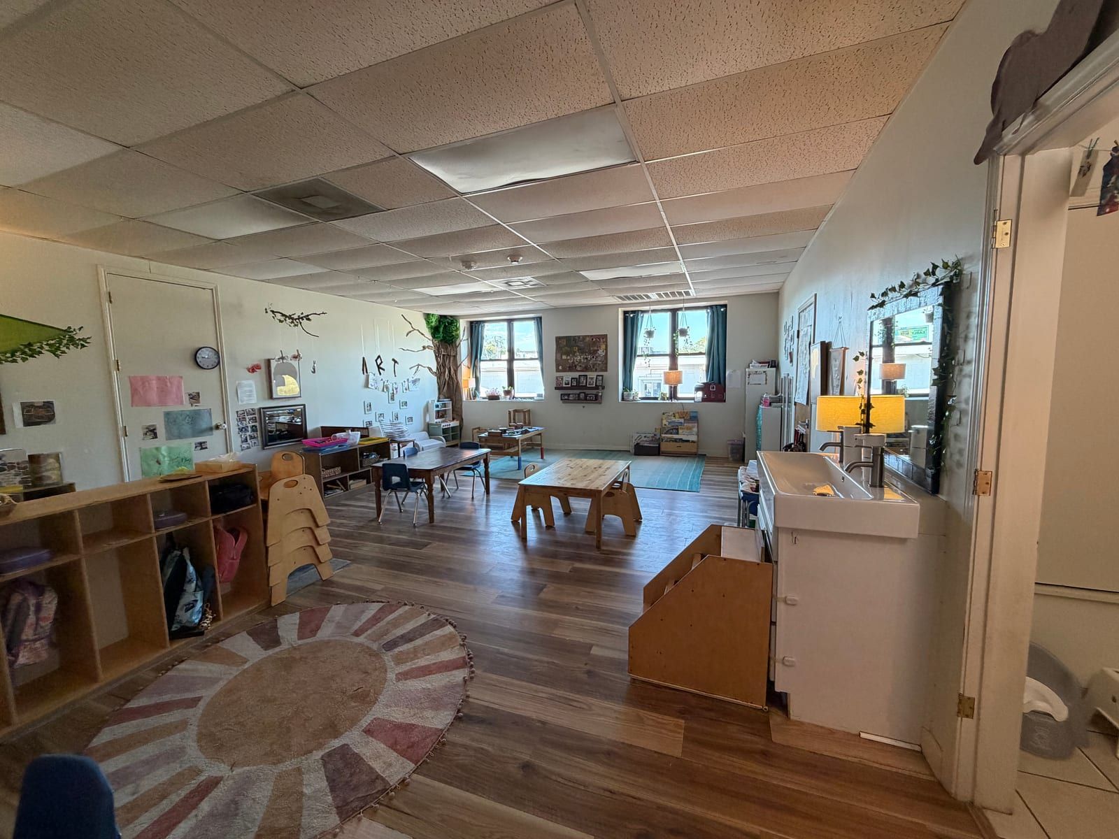 A long classroom with a round sun-patterned rug, cubbies by the door, low tables and windows along the far wall.