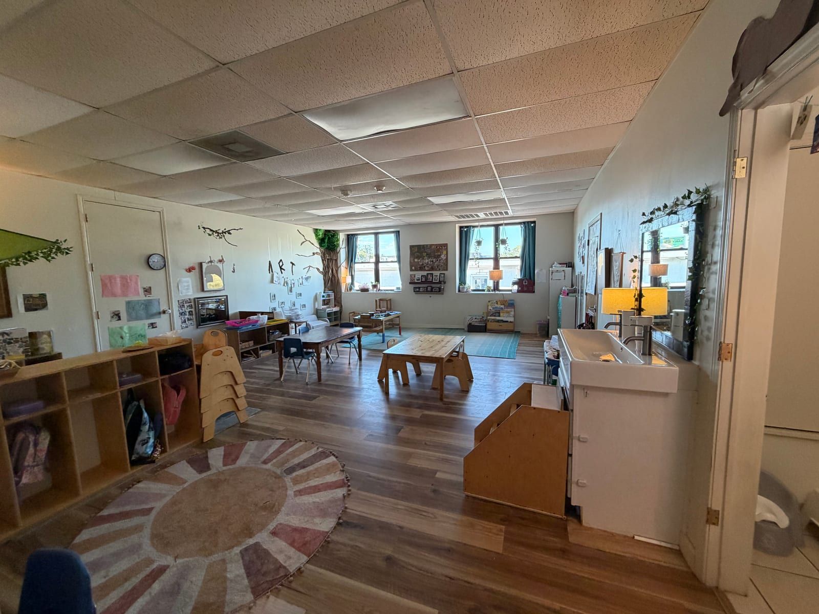 A classroom with a round sun-patterned rug, cubbies, small wooden tables and windows along the far wall.