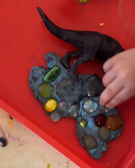 A child's fingers press glass gems into a slab of blue-grey dough with a toy dinosaur on top, in a red tray.