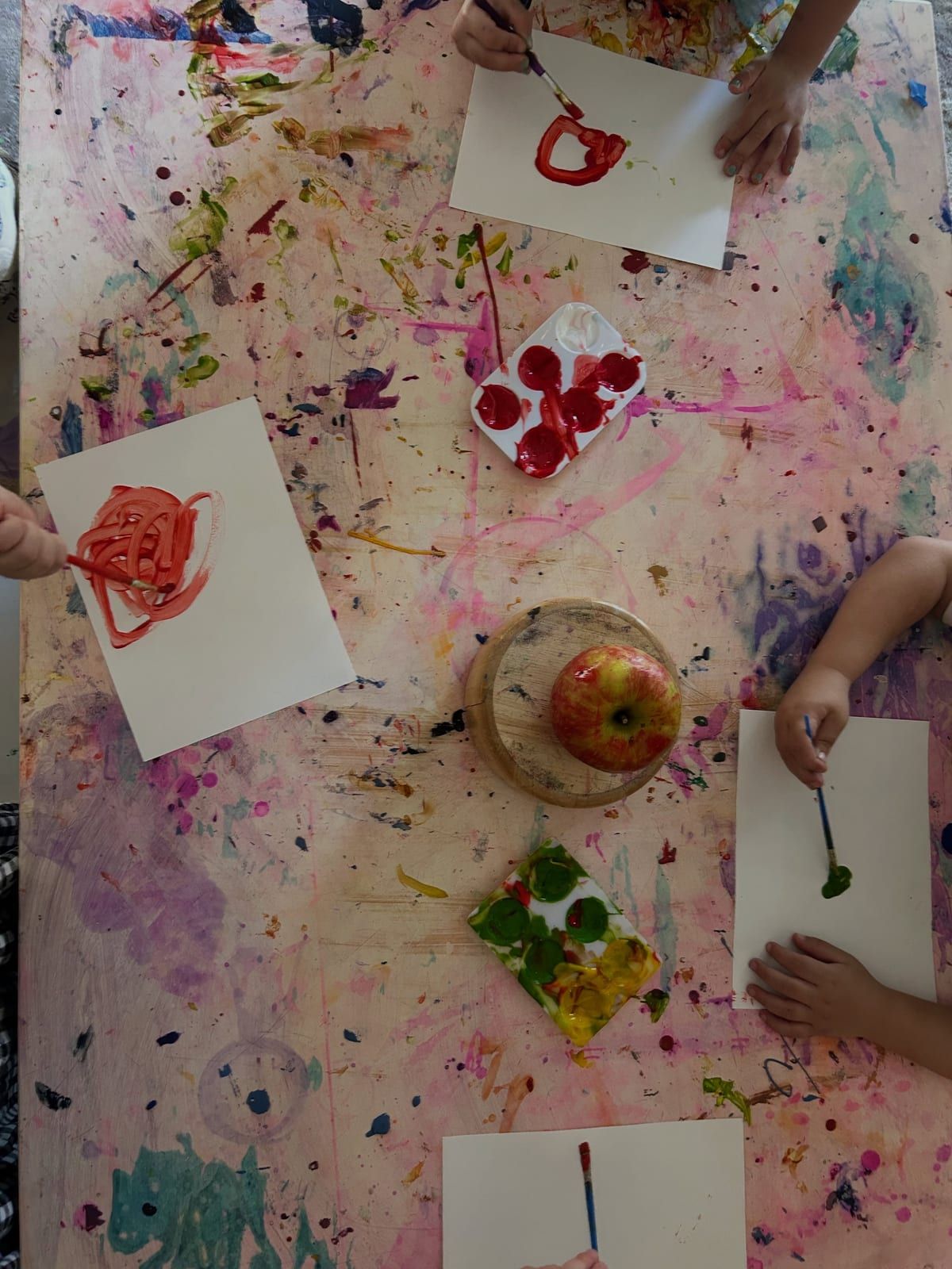 Children painting at a paint-spattered table around an apple set out for a still life.