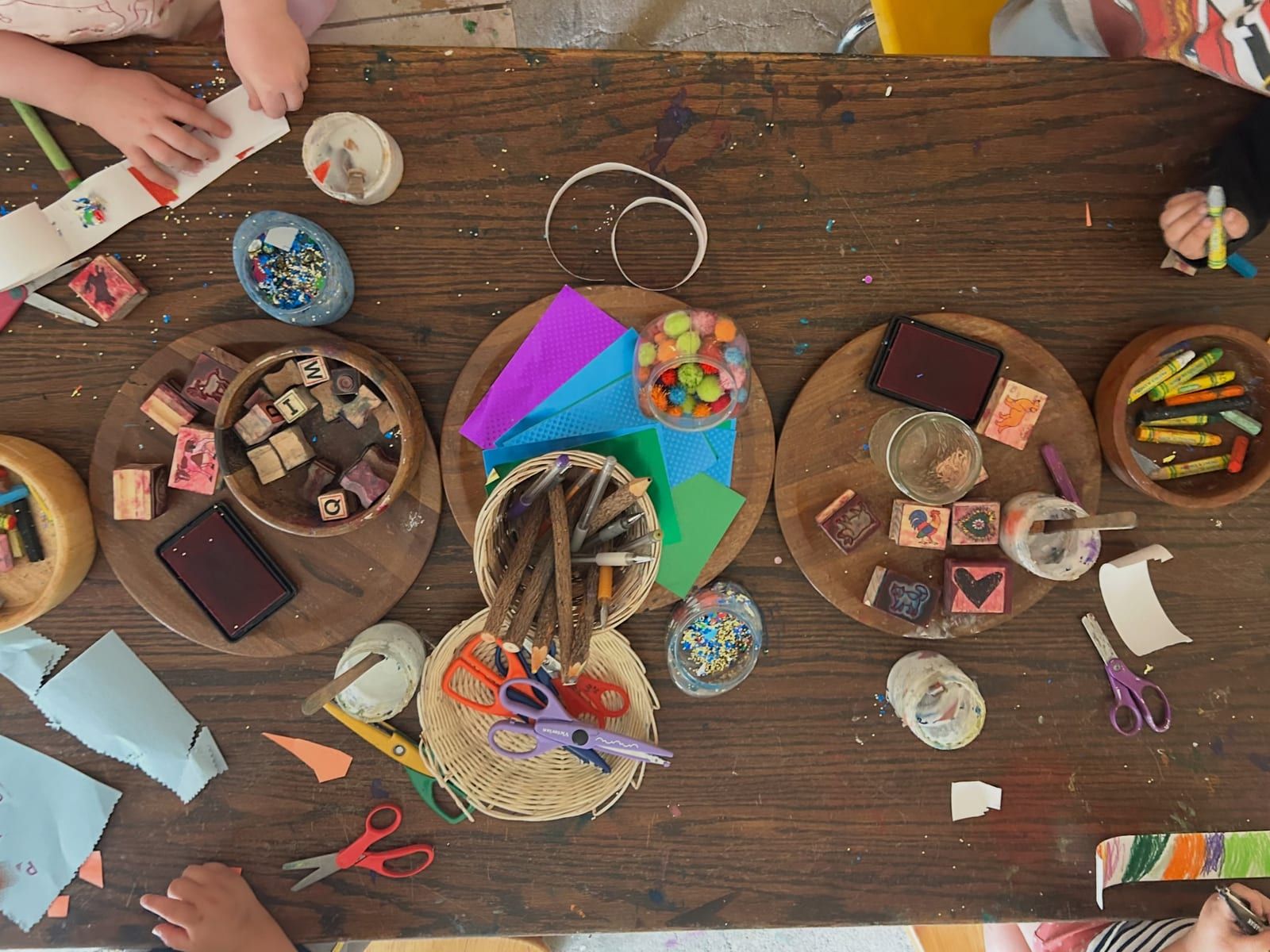 A wooden table set with trays of art materials: tissue paper, stamps, ink pads, beads, sticks and scissors.