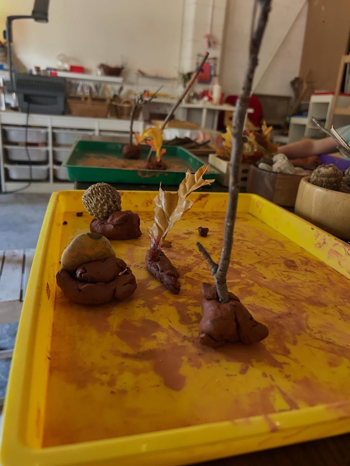 Dough sculptures with twigs, acorns and leaves on a yellow tray, with classroom shelves behind.
