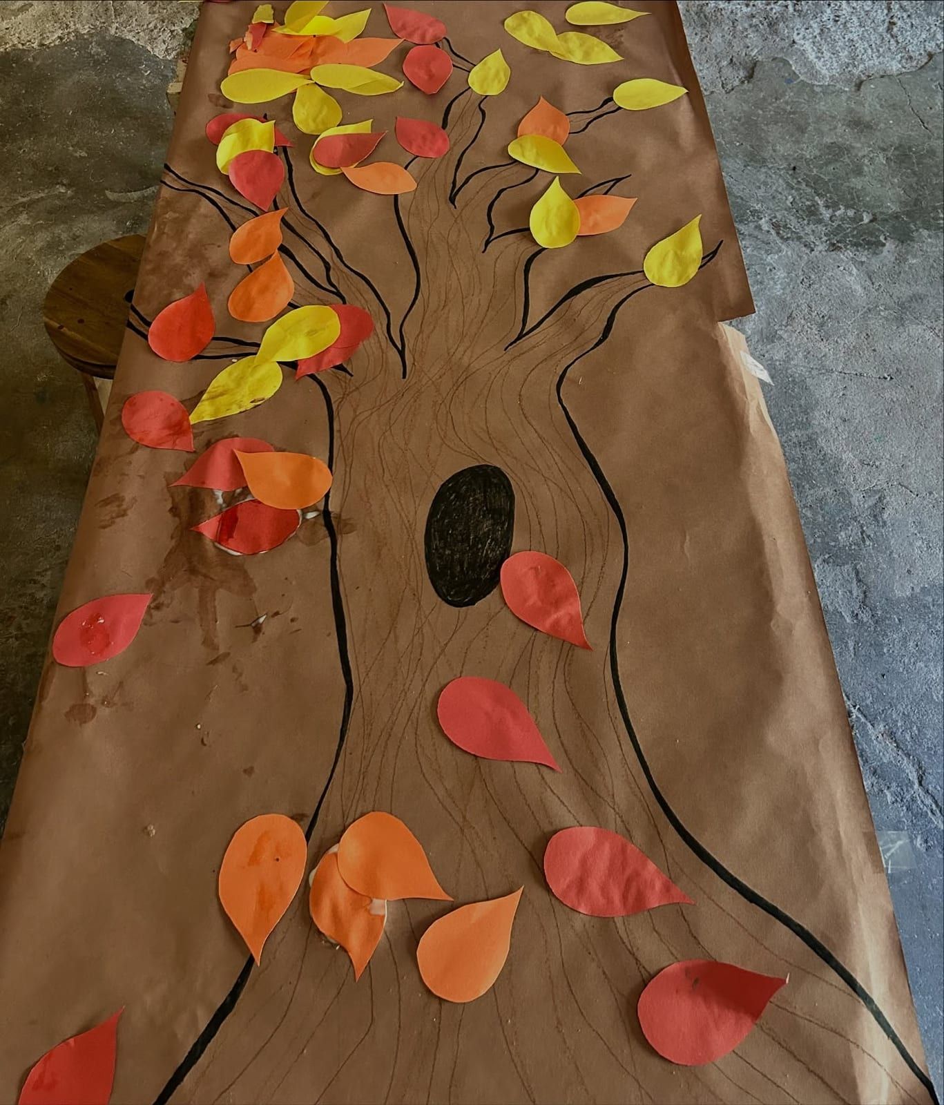 A tree trunk drawn on brown paper, covered in red, orange and yellow paper leaves.