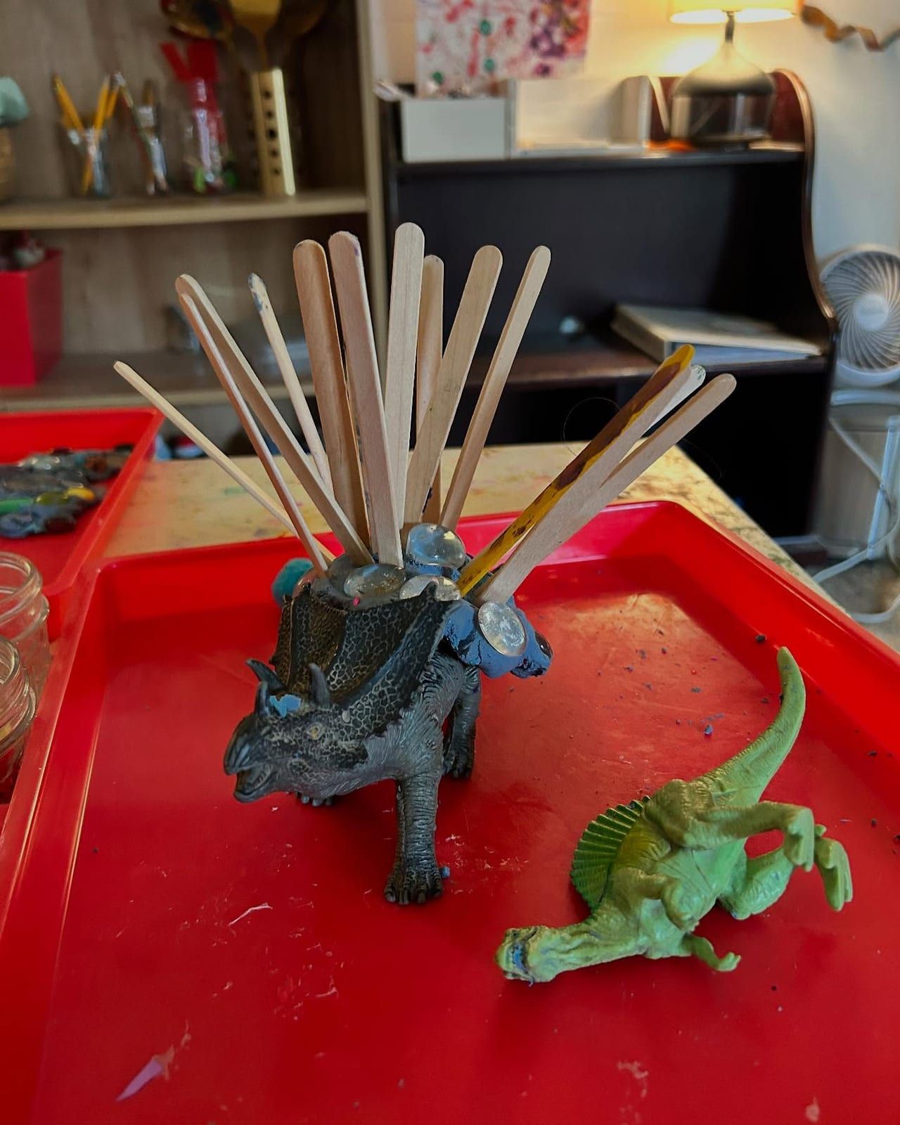 A toy dinosaur bristling with craft sticks on a red tray, with a classroom shelf behind.