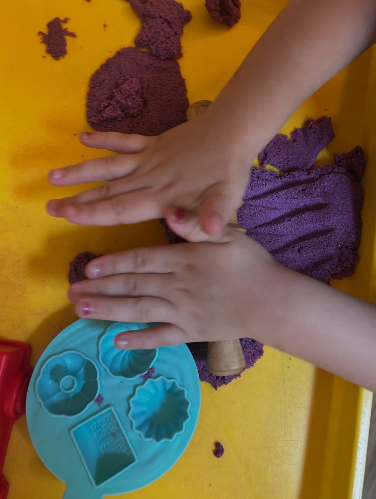 Two hands pressing purple dough beside a blue mold of flower and shape cutouts, on a yellow tray.