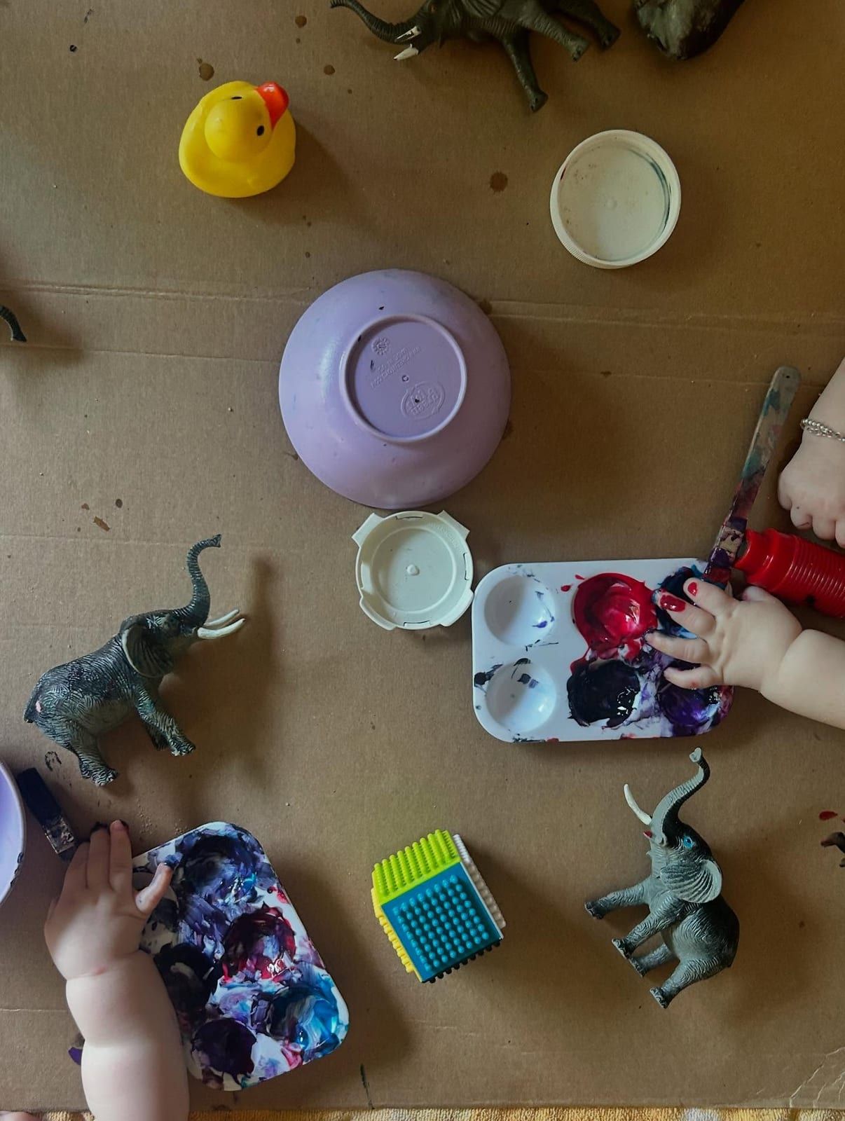 Little hands painting beside a paint palette, a purple bowl, toy elephants and a rubber duck on cardboard.