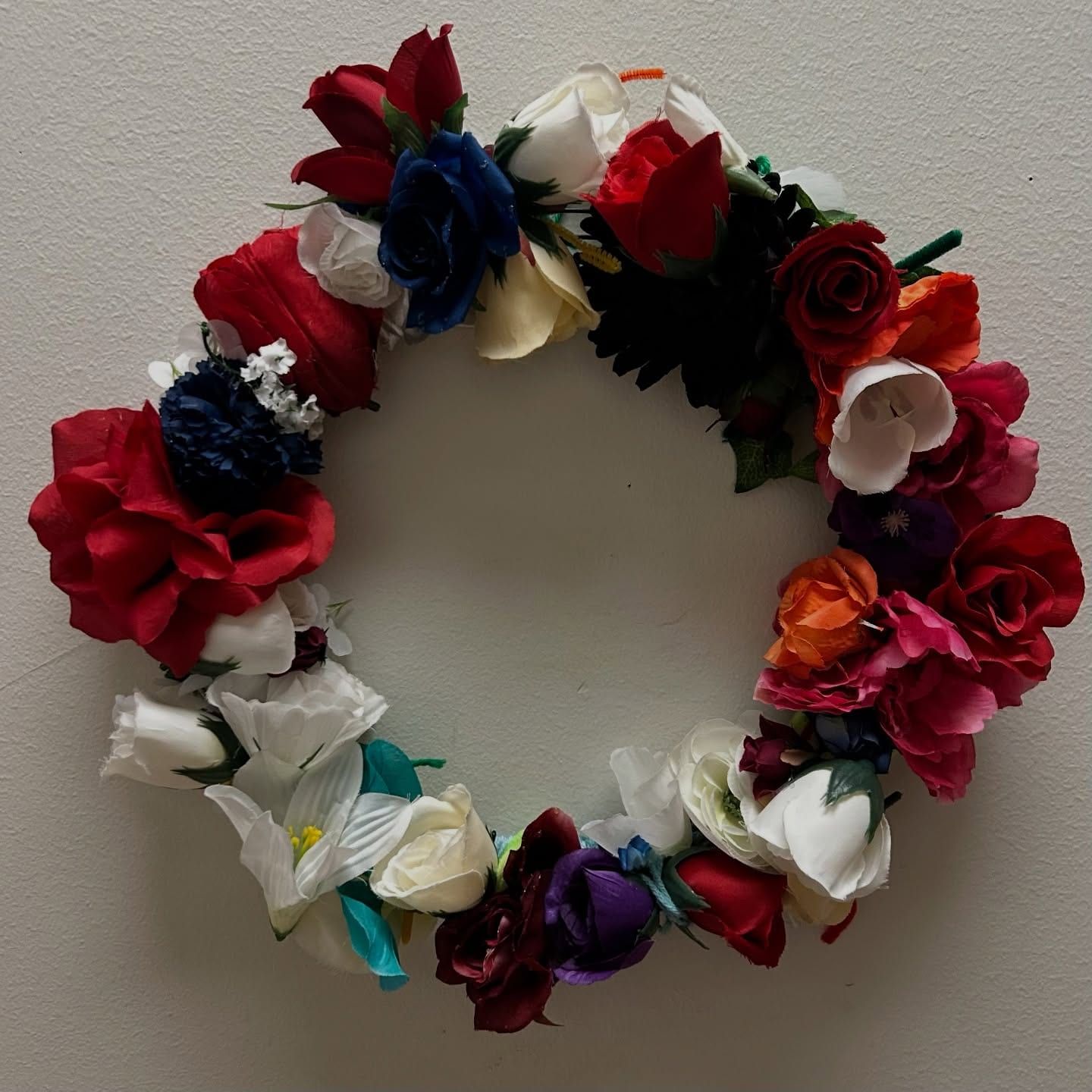 A finished wreath of red, white, blue and orange fabric flowers hung on a white wall.