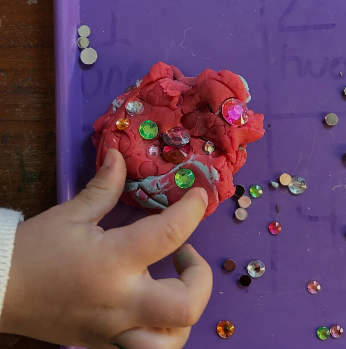 A child's fingers pressing gems and sequins into a ball of red dough on a purple tray.