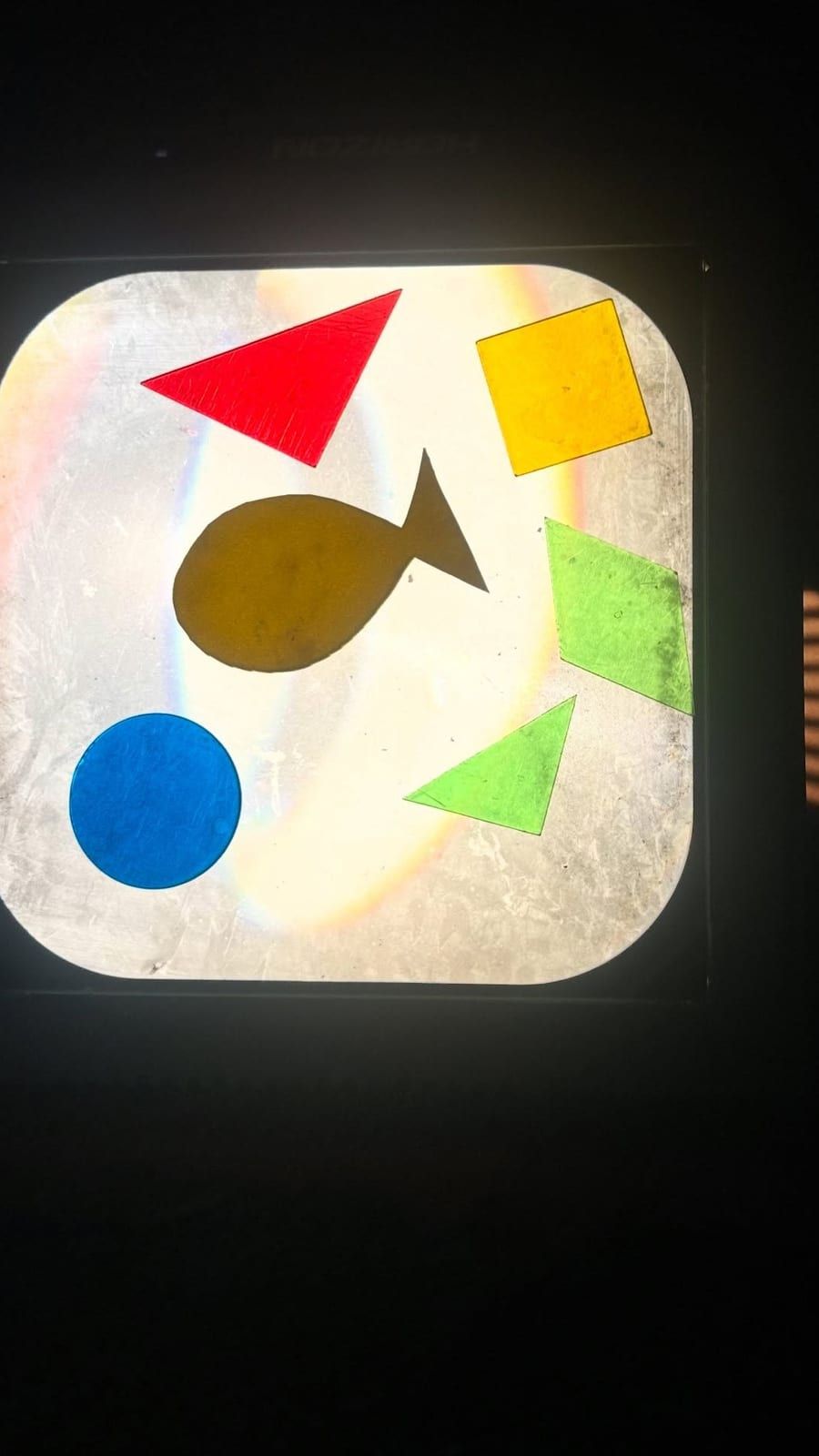 A glowing light table holding cut shapes in colored film: a red triangle, a yellow square, a blue circle, green triangles and a fish.