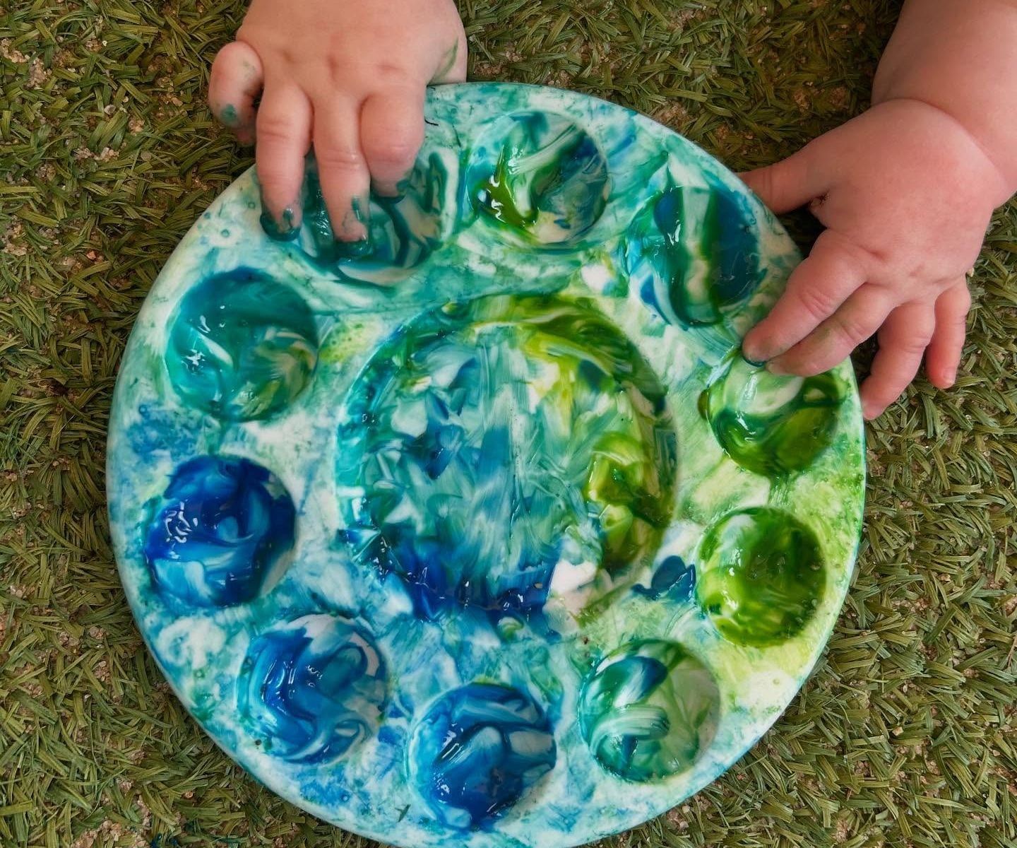 Two small hands resting on a round paint palette swirled with blue and green, on artificial grass.