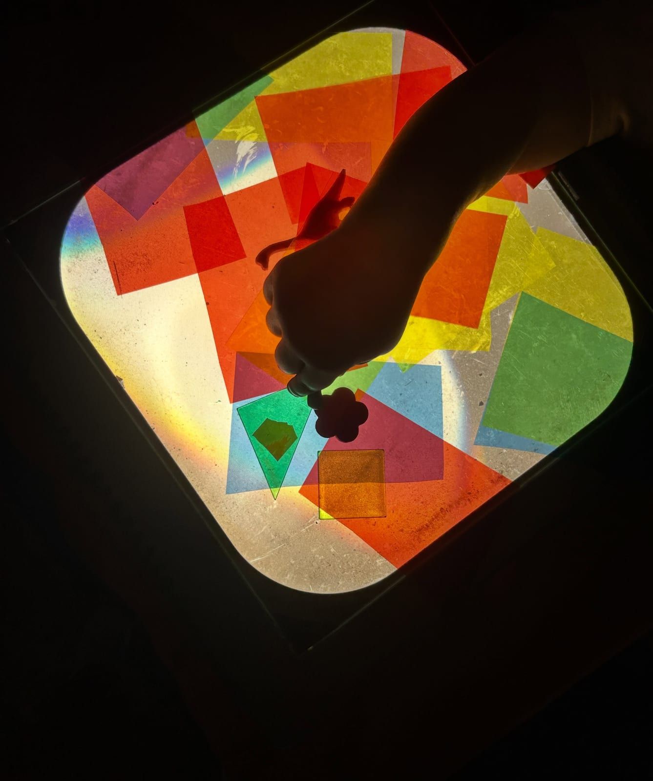 A child's hand arranging squares of colored tissue paper on a glowing light table.