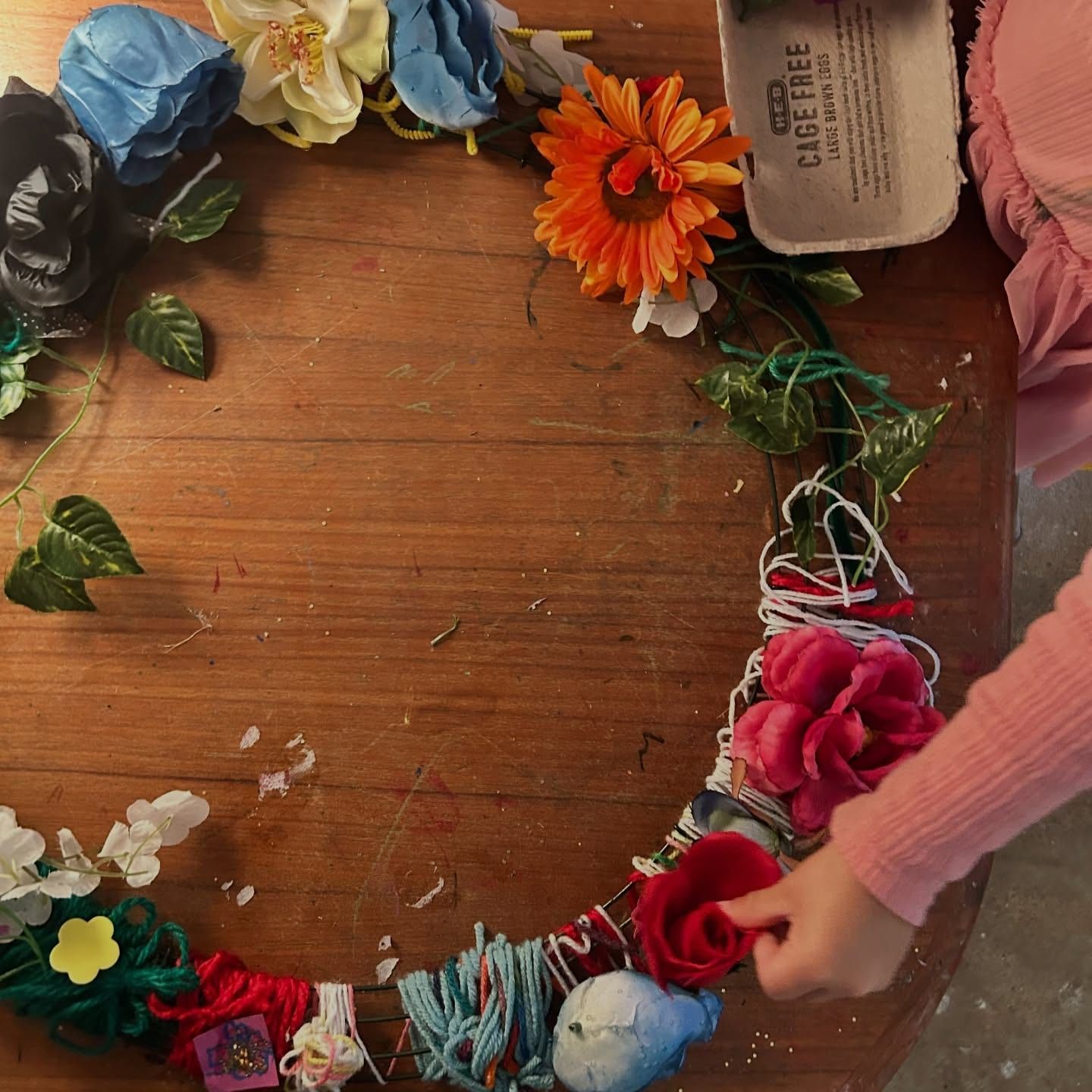 Hands tucking fabric flowers and yarn into a wreath frame on a wooden table.