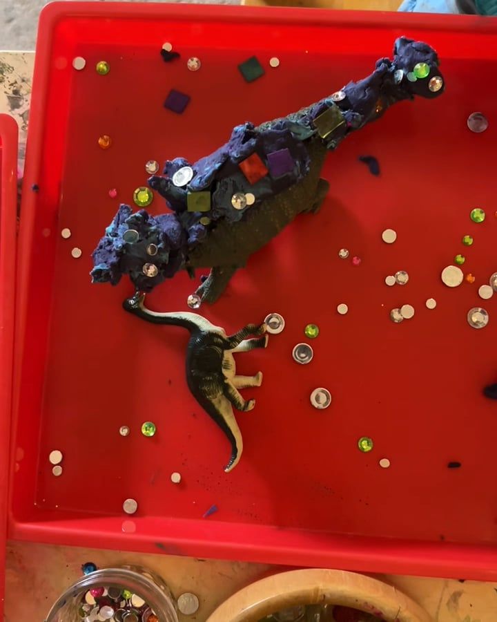 A toy dinosaur coated in purple dough and studded with sequins and plastic gems, in a red tray scattered with more gems.