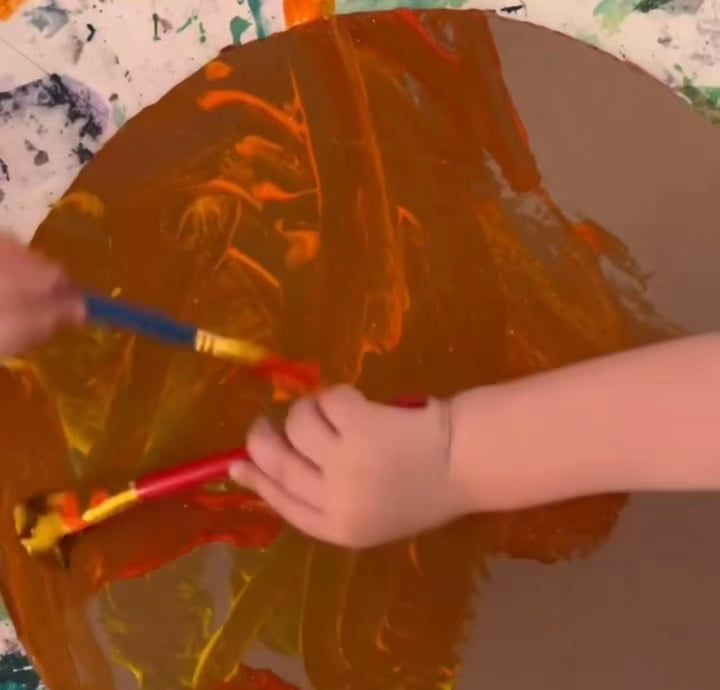 Two children's hands with brushes spread orange and yellow paint across a large cardboard circle.