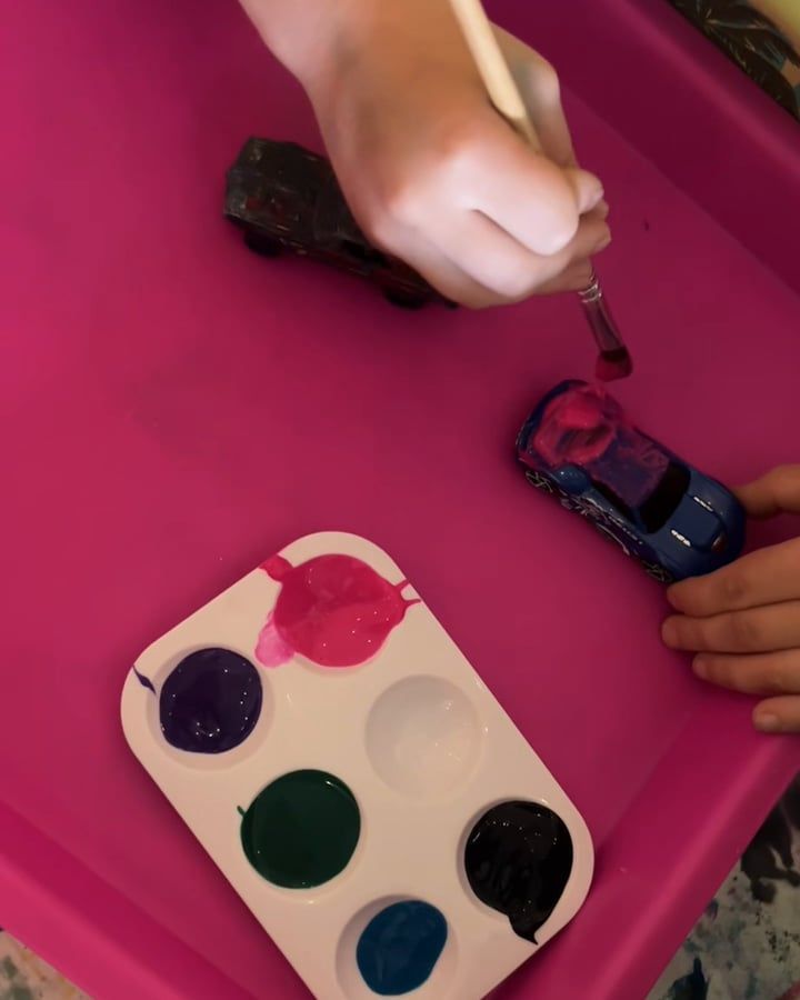 A child's hand brushes pink paint onto a blue toy car in a pink tray, beside a palette of purple, pink, green, black and blue paint.