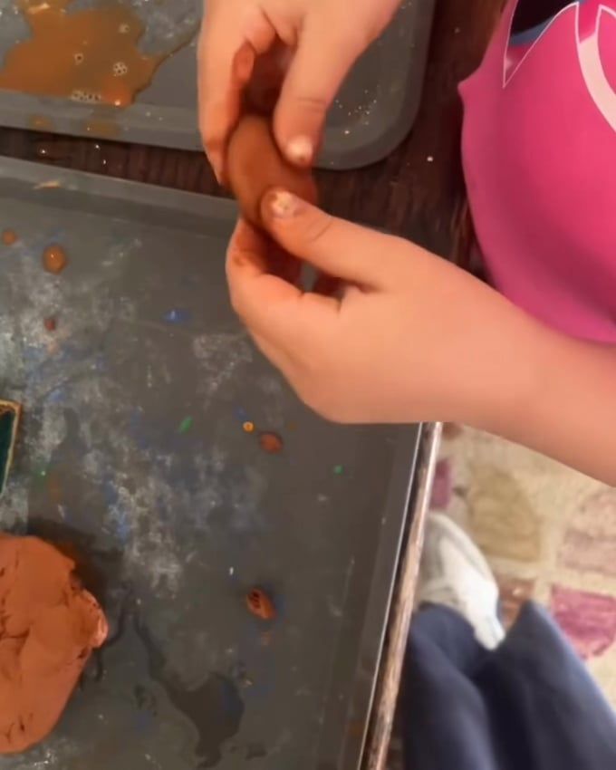 A child's hands roll a small piece of reddish-brown clay above a metal tray.