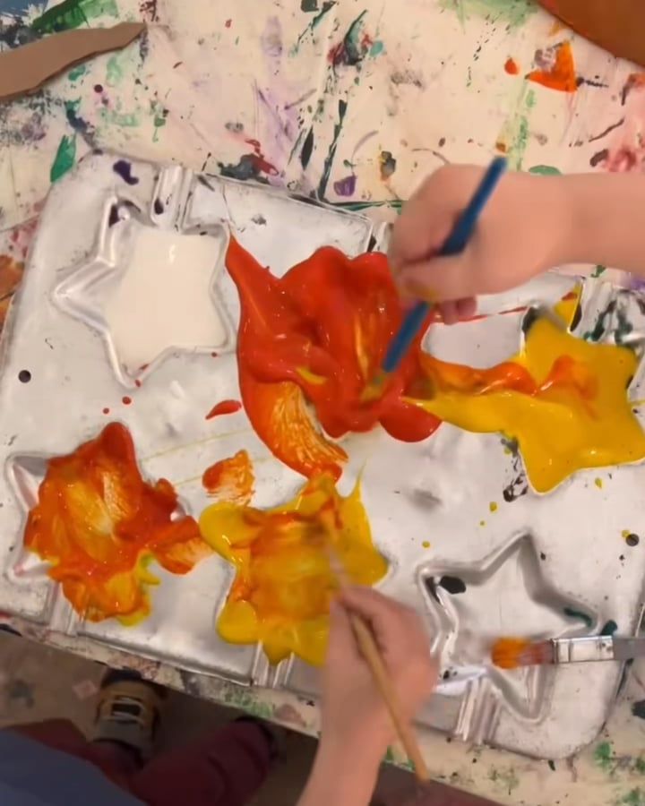 Two children's hands with brushes swirl red, orange and yellow paint in the star-shaped cups of a metal tray.