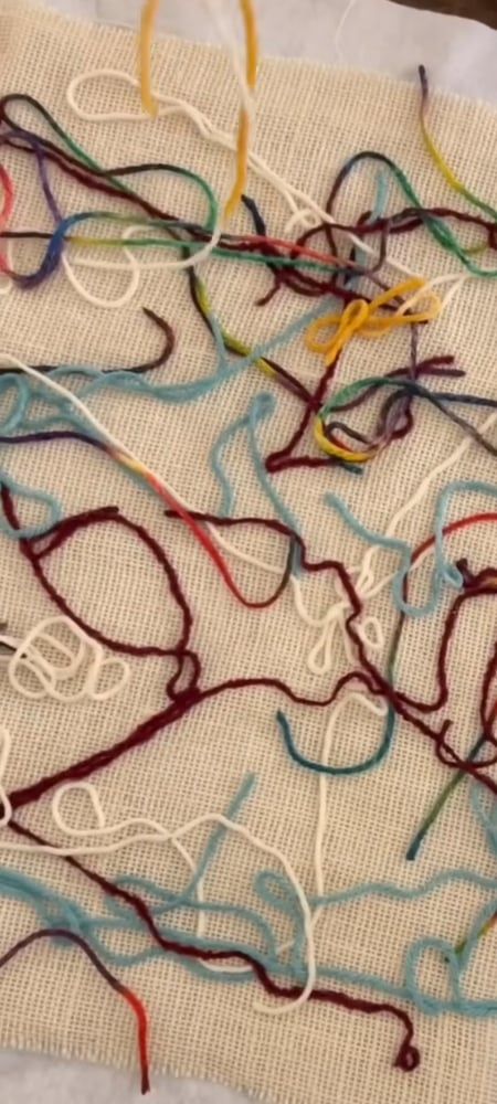 Loops of maroon, sky-blue, white and rainbow yarn laid across a square of loosely woven cloth.